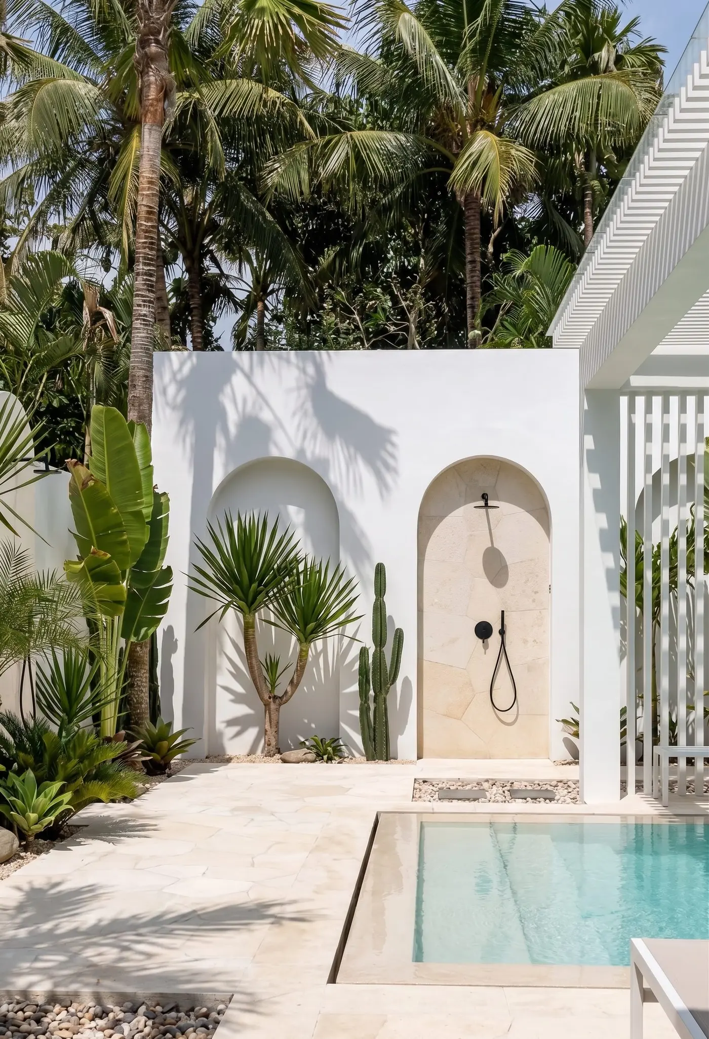 White-rendered courtyard with twin arches, dragon tree and column cactus, black-tapware shower under the right arch, crazy-paved pale stone, aqua pool and a palm canopy overhead